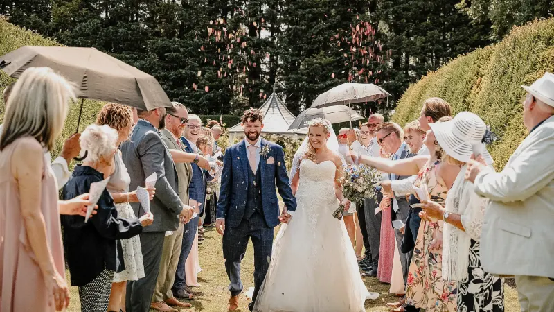 Bride and groom walking between wedding guests throwing confetti in a garden.